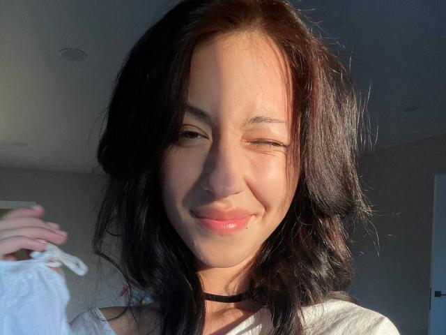 Maryln Werntz, com cabelo preto ondulado, sorrindo com um olho fechado, lábios rosados brilhantes, camisa branca, colar preto, segurando um tecido branco, com luz do sol no lado direito do rosto..