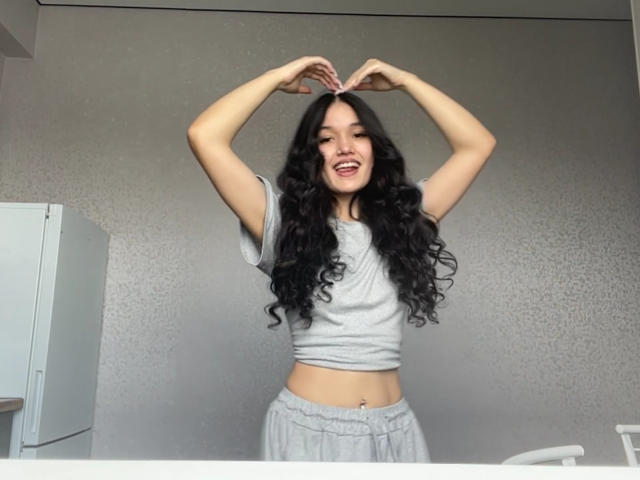 Meaghan Grigalonis mit langen, lockigen, schwarzen Haaren, einem grauen Crop-Top und grauen Pyjamahosen, lächelnd, mit den Händen, die über ihrem Kopf ein Herz formen, steht in einem Raum..