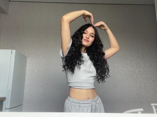 Meaghan Grigalonis mit langen, lockigen, schwarzen Haaren, einem grauen Crop-Top und grauen Hosen, mit erhobenen Armen, die einen Bauchnabelpiercing zeigt, lächelnd..