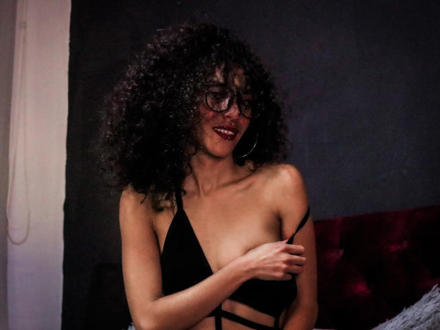 LiliMuller, com cabelos pretos cacheados, óculos e batom vermelho, vestindo uma blusa preta com alças finas, cobrindo levemente os seios com a mão, sorrindo de forma sedutora..