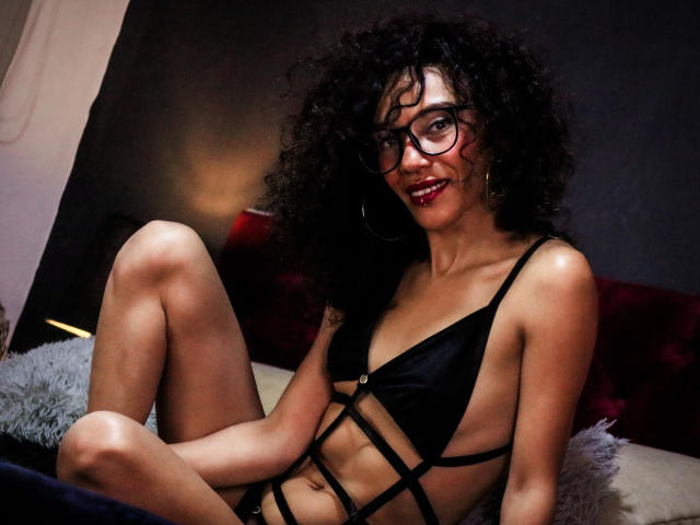 LiliMuller, con cabello negro rizado, usando gafas y un conjunto de lencería negra con tirantes finos, sentada con las piernas levantadas, sonriendo, mostrando lápiz labial rojo y pequeños pechos firmes..