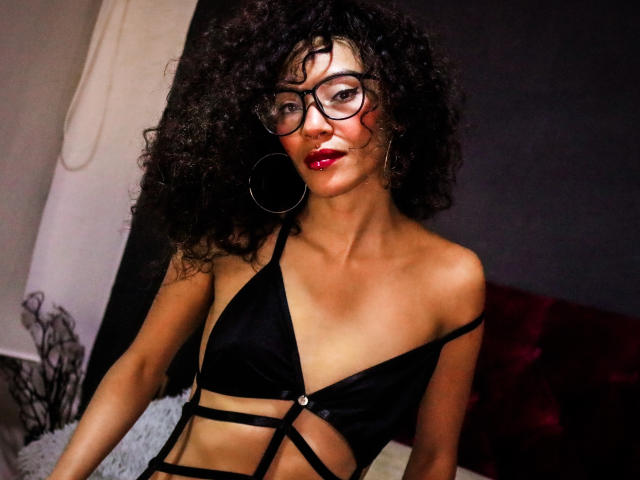 Lili Muller, com cabelos pretos cacheados, batom vermelho, óculos, brincos grandes e um sutiã preto com alças finas, mostrando seus seios e abdômen tonificado, aparentando ser sexy e confiante..