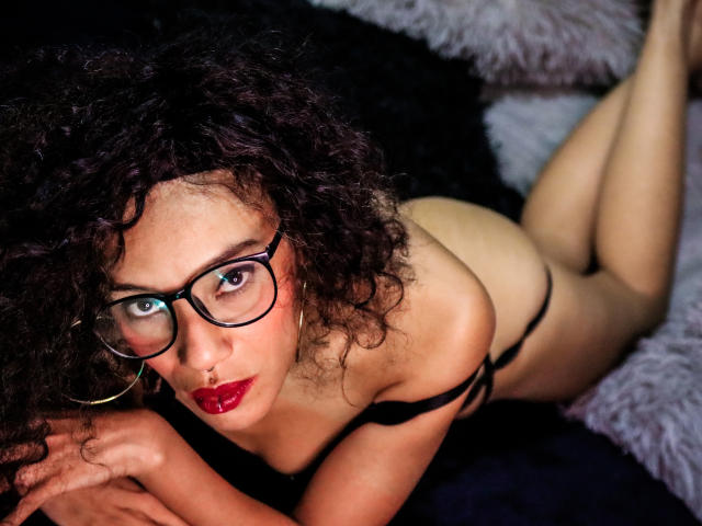 LiliMuller, con cabello negro rizado, gafas, lápiz labial rojo, un sujetador negro y una tanga, está acostada sobre una superficie negra y gris con pelusa, mirando hacia arriba con una expresión seria, mostrando sus pechos..