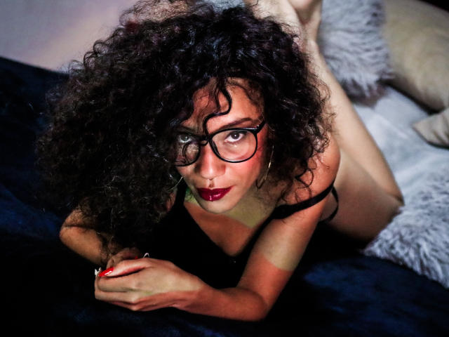LiliMuller, avec ses cheveux noirs bouclés, son rouge à lèvres rouge, ses lunettes et sa lingerie noire, est allongée sur un lit bleu foncé, regardant de manière séduisante l'appareil photo, les jambes pliées derrière elle..