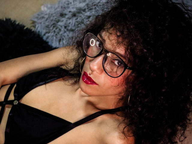 LiliMuller, con cabello negro rizado, usando gafas, lápiz labial rojo y un top negro con tirantes, mirando hacia arriba con una ligera sonrisa. Sus senos son visibles, mostrando escote..
