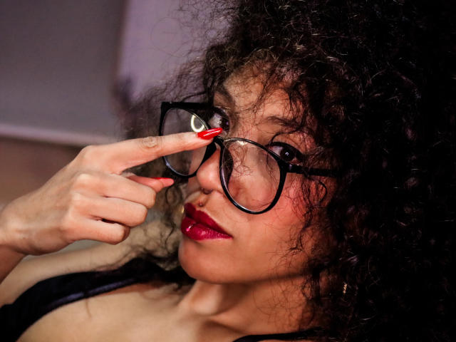 LiliMuller, com cabelos pretos encaracolados, usando óculos pretos, batom vermelho e unhas vermelhas, tocando seus óculos com o dedo indicador, olhando ligeiramente para o lado..