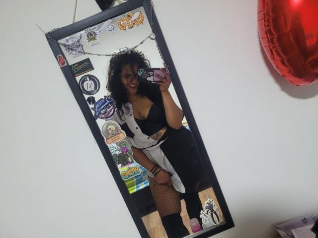 ZiaNoir with curly black hair, smiling, wearing a black bra, white jacket, black skirt, fishnet stockings, and black boots, taking a mirror selfie, showing her tits and ass.