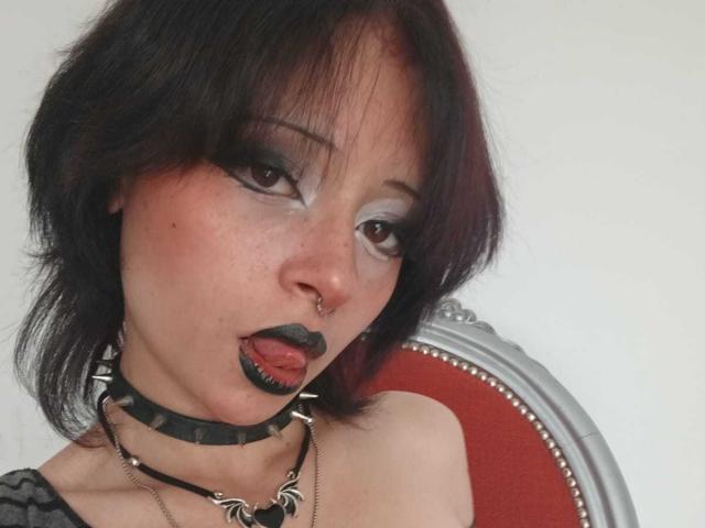 ViolettClay has dark red hair, heavy black eyeliner, black lipstick, nose piercing, black choker with spikes, and tongue out. Her tits are slightly visible. She's wearing a grey top.