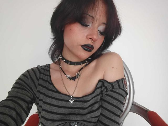 ViolettClay tem batom preto, um piercing no nariz e maquiagem escura nos olhos. Ela está vestindo uma camisa listrada, um colar e um colar com um pentagrama. Ela está sentada em uma cadeira..