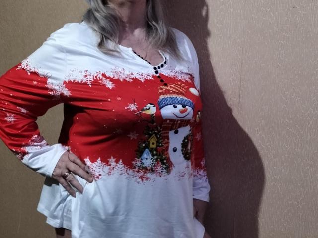 LolkaAlyss, avec ses cheveux gris, porte une chemise de Noël rouge et blanche représentant un bonhomme de neige, et se tient debout, une main sur la hanche. La chemise est ornée de flocons de neige, d'un bouton et d'un motif hivernal décoratif..