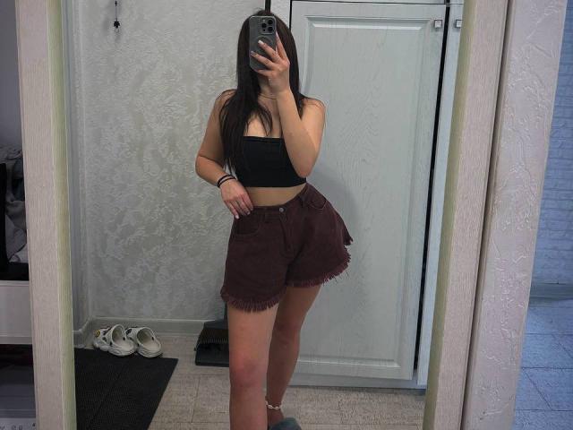 InesGueningsman with long black hair, black tube top, and frayed burgundy shorts, taking a mirror selfie, showing her tits and ass, holding a phone, in a room.