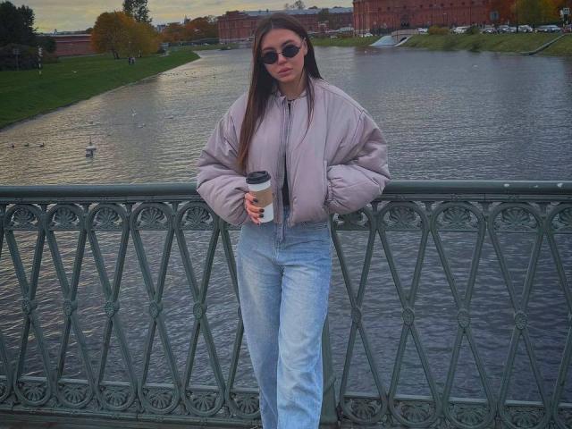 InesGueningsman with long brown hair, wearing sunglasses, a light purple jacket, and blue jeans, holding a coffee cup, leaning on a green iron bridge.