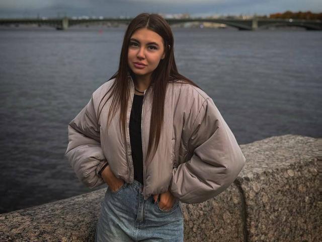 InesGueningsman with long brown hair, hands in jeans, wearing a puffy beige jacket, standing by a stone wall, looking slightly to the side, overcast sky, calm water.