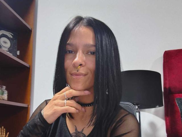 AmelieBlake with black hair, nose ring, choker, and sheer top, smiling, hand on chin, tattoo on chest, black lamp in background, red chair partially visible.