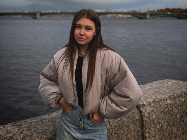 InesGueningsman with long brown hair, hands in jeans pockets, wearing a puffy beige jacket, standing against a stone wall, looking at the camera with a slight smile.