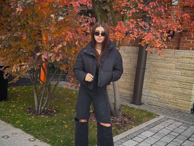 InesGueningsman has long brown hair, wears black sunglasses, a puffy black jacket, and ripped black jeans. Autumn tree with red leaves is behind her. She stands on a brick path.