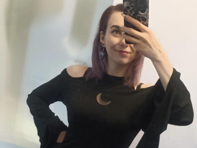 VelvetTanne with purple hair, smiling, taking a mirror selfie in a black off-shoulder top with a crescent moon design, showing her tits, holding a phone with a black case.