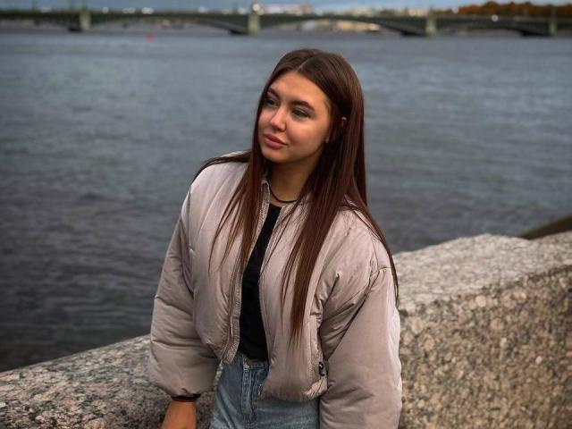InesGueningsman with long brown hair, wearing a puffy beige jacket and blue jeans, standing by a stone wall, looking slightly to the side, with a calm water background.