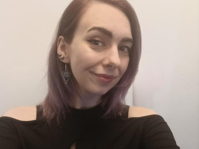 VelvetTanne, con i capelli tinti di viola, indossa una maglietta nera a spalle scoperte, ha il rossetto rosso, sorride, porta orecchini geometrici e un anello al naso, e guarda verso la fotocamera..