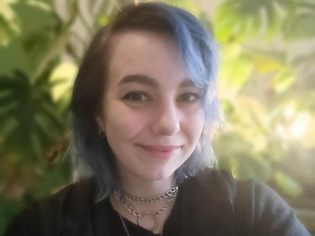 VelvetTanne, con el cabello teñido de azul, un aro en la nariz, una blusa negra y collares de cadena, sonríe suavemente. Sus pechos son pequeños y tiene una apariencia linda, ligeramente traviesa..