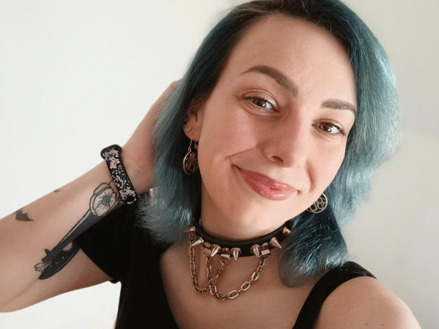 VelvetTanne with blue-dyed hair, black top, and chain collar, smiling with tongue out, has tattoos, ear rings, and a bracelet, looking cute and sexy.