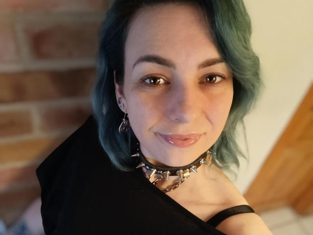 VelvetTanne has teal hair, a black choker with metal spikes, and a black top, smiling with glossy pink lips, brown eyes, and small earrings, looking up at the camera.