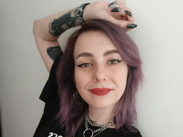 VelvetTanne mit lila Haaren, schwarzem Eyeliner, rotem Lippenstift, schwarzen Nägeln, Tattoos an den Armen, einer Kettenkette, einem schwarzen T-Shirt, mit der Hand über dem Kopf, lächelnd, blasser Haut und braunen Augen..
