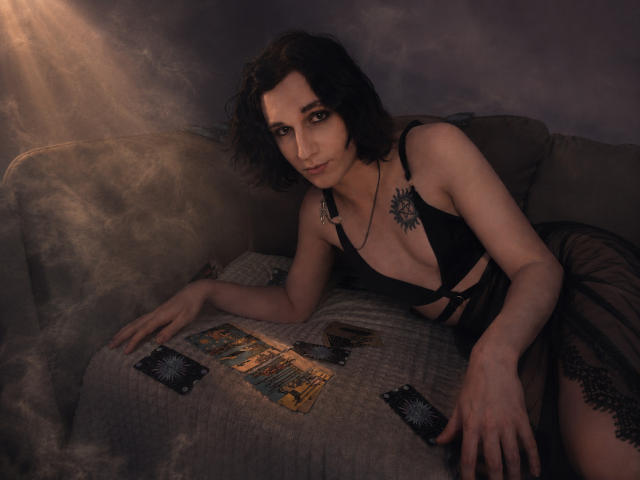 ReginaMay, com cabelos cacheados escuros, lingerie de renda preta, raios de sol e tatuagens, deitada em um sofá cinza, mostrando seios pequenos, e algumas cartas sobre um cobertor..