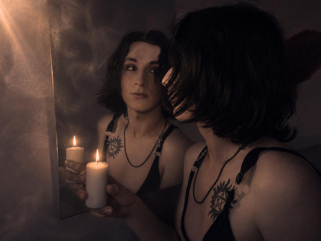 ReginaMay com cabelo preto curto, sutiã de renda preta, tatuagem no peito, segurando velas, olhando para o reflexo, quarto escuro e esfumaçado, luz de velas suave, expressão concentrada, sombras sutis, clima íntimo..