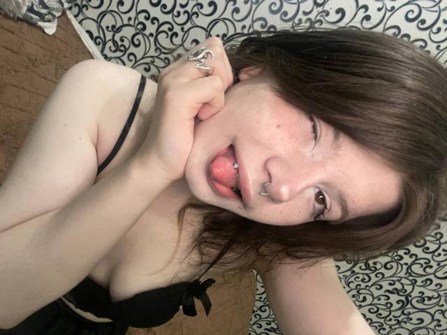 Marla Rotan heeft bleke huid, bruin haar en een neusring. Ze steekt haar tong uit met een tongpiercing, draagt zwarte lingerie en raakt haar wang aan..
