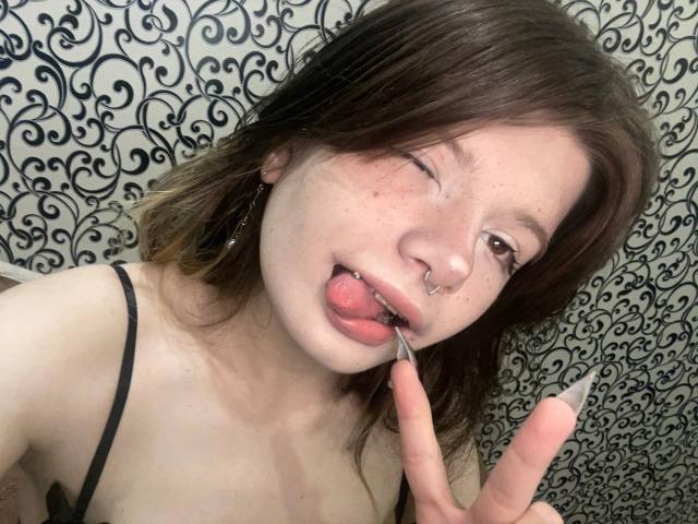 Marla Rotan, avec ses cheveux bruns, son anneau de nez et ses taches de rousseur, tirant la langue, léchant une brosse à dents, faisant un signe de paix, portant un haut noir à bretelles fines, clignant de l'œil..