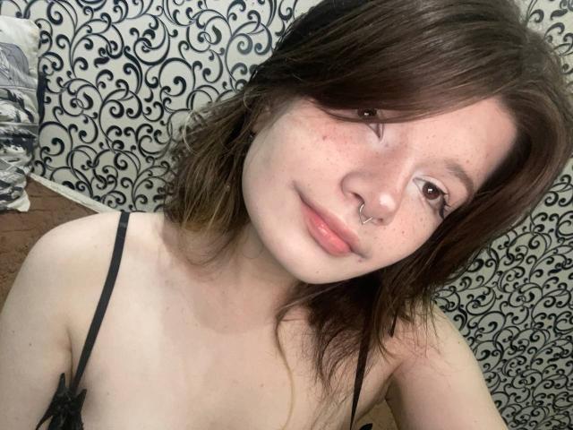 Marla Rotan, met sproeten, een neusring en bruin haar, glimlacht lichtjes en laat kleine borsten zien in een zwart topje met bandjes, terwijl ze een selfie maakt..
