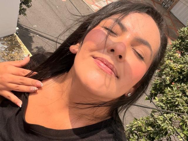 AntonelaVega with black hair, sunlit face, pink blush, eyes closed, black shirt, hand on neck, slight smile, shiny skin, pink lips, small diamond earring, sunlight, greenery.
