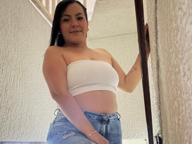 Antonela Vega, met zwart haar, lachend, draagt een witte top zonder bandjes, waarbij haar borsten zichtbaar zijn, en blauwe jeans, staat in een deuropening en houdt een deurkozijn vast..