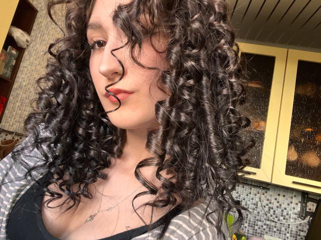 EricParsygnat a les cheveux noirs et bouclés, une peau pâle, et porte une chemise rayée avec un soutien-gorge noir, laissant entrevoir une partie de sa poitrine. Elle regarde l'appareil photo avec une légère moue..