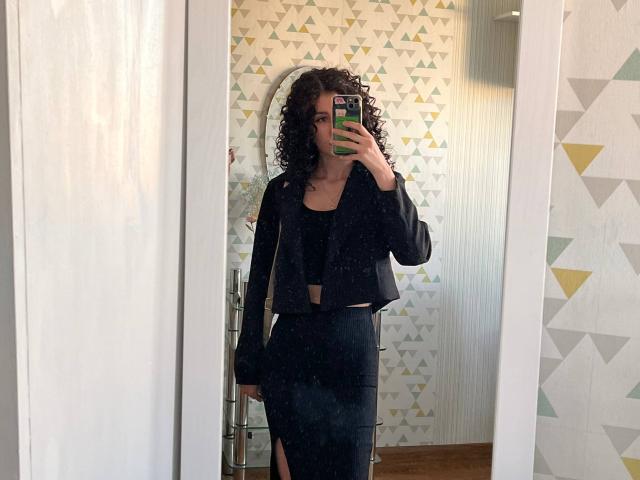 Eric Parsygnat, con cabello negro rizado, vistiendo una chaqueta corta negra y una falda negra con una abertura en el muslo, tomando una selfie frente al espejo con una funda de teléfono verde..