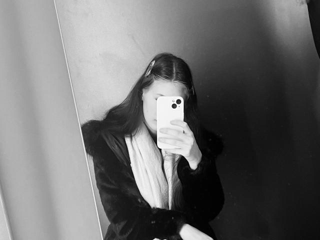 Eric Parsygnat, mit langem, dunklem Haar, trägt einen dunklen Pelzmantel und einen weißen Schal und macht ein Selfie mit seinem Smartphone, wobei sein Gesicht verdeckt ist. Schwarz-weiß-Bild..
