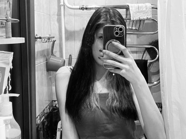 Eric Parsygnat, avec de longs cheveux foncés, prend un selfie devant un miroir, tenant un téléphone, portant un haut sans manches, avec des seins visibles, des ongles noirs, dans une salle de bain, photo en noir et blanc, reflet dans le miroir..