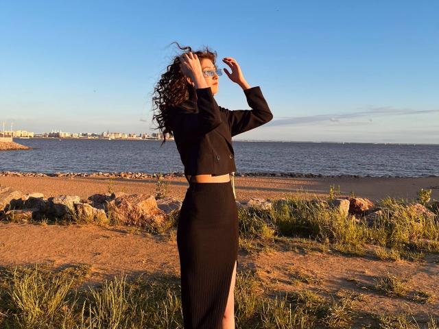 Eric Parsygnat, avec ses cheveux bruns bouclés, portant un crop top noir et une jupe noire taille haute, se tient sur une plage rocheuse, les mains dans les cheveux, sous un ciel bleu clair, au coucher du soleil..