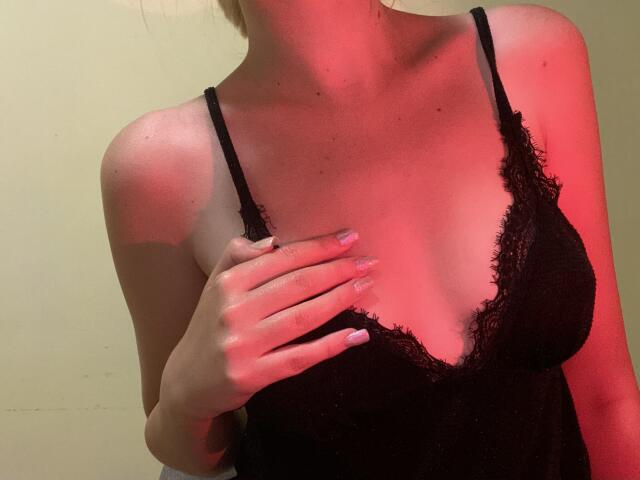 FlyFreedom with light skin, wearing a black lace camisole, has a hand on her tits, pink-nail-polished fingers, under red lighting, showing her cleavage and slim shoulders.