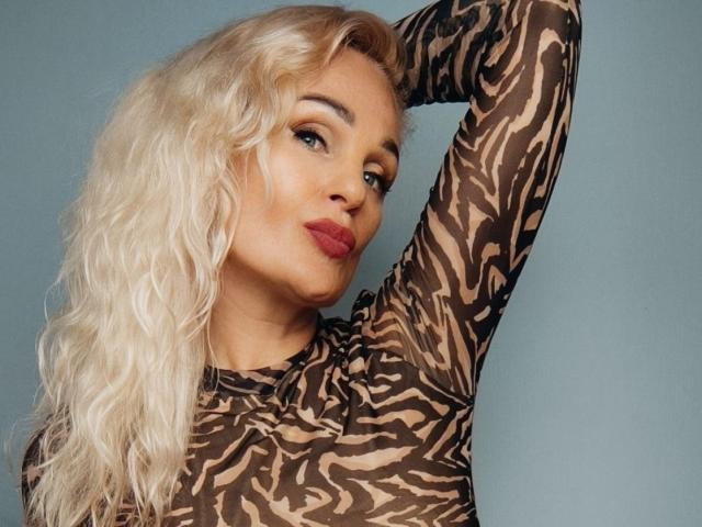 MiaDreamy, avec ses cheveux blonds ondulés, portant un haut à rayures zébrées, a le bras levé, montrant ses seins, avec du rouge à lèvres et une expression confiante..