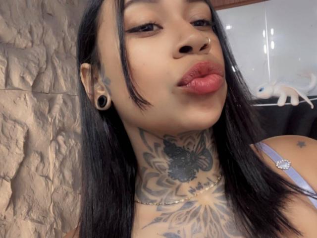 HollyCattaleiya ha i capelli neri, un piercing al naso, grandi espansori alle orecchie neri e diversi tatuaggi sul collo e sul petto. Ha le labbra corrugate e indossa una maglietta viola..