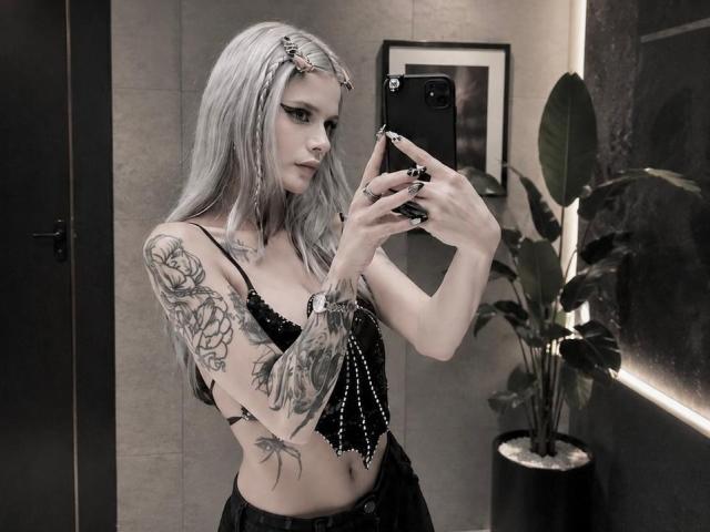 ErgoProxy com cabelo prateado, tatuagens, blusa de renda preta, segurando um telefone, selfie no espelho, tatuagens visíveis no braço e na lateral, calças pretas, joias, expressão séria, em um quarto com pouca luz..