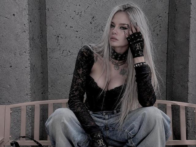 ErgoProxy com cabelo loiro comprido, blusa de renda preta, jeans, luvas pretas e colar, sentada em um banco rosa, com a mão no rosto, com uma expressão séria, com tatuagens visíveis, e um fundo de parede cinza..