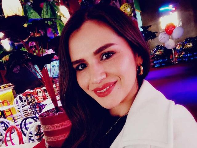 CameronViviane, com longos cabelos castanhos, sorrindo com batom rosa, vestindo um casaco branco, tem um rosto redondo e fofo e um nariz pequeno, e está em uma loja colorida e bem iluminada..