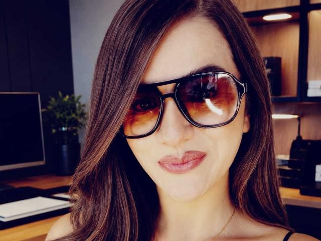 Cameron, Viviane, com cabelos castanhos ondulados, usando óculos de sol retangulares pretos e um sorriso discreto, tem lábios cheios e está olhando diretamente para a câmera..