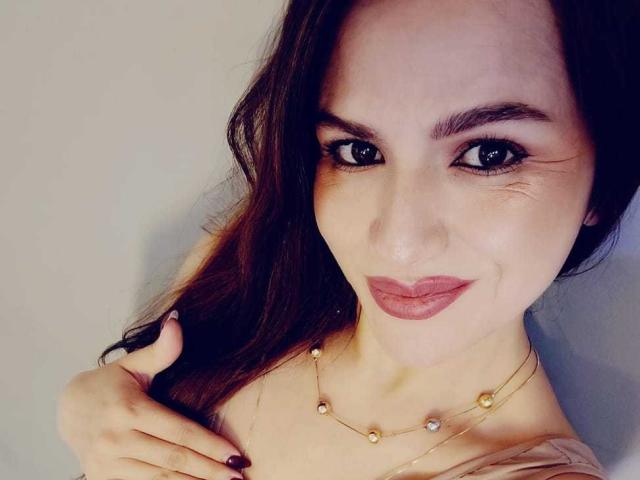CameronViviane with long brown hair, dark eyes, and pink lipstick, wearing a gold necklace, touching her hair with purple-nailed fingers, smiling slightly.