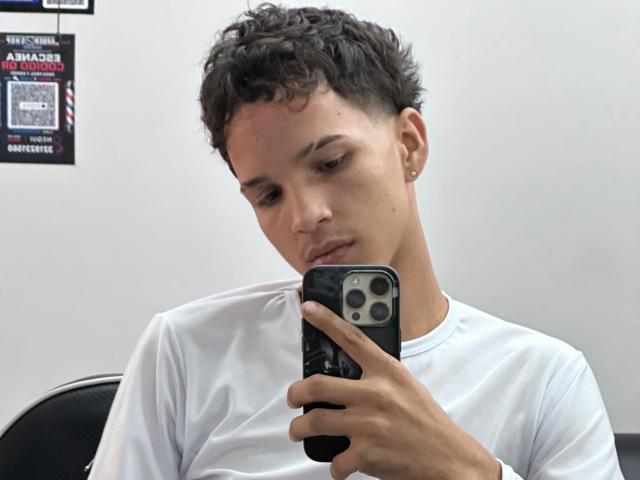 MidnightKing, avec ses courts cheveux bruns bouclés, sa chemise blanche et ses petites boucles d'oreilles, regarde un téléphone noir équipé de trois caméras. Sa peau est claire et il est assis sur une chaise..