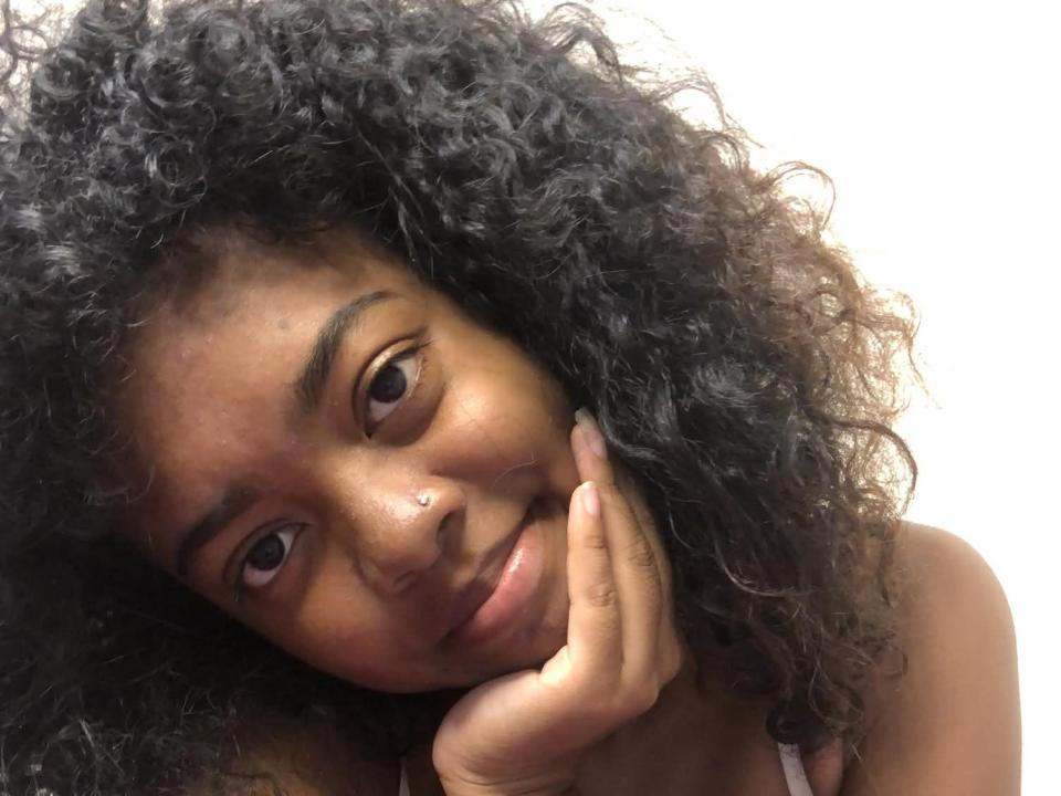 SweetDollSexy, com cabelo preto encaracolado, pele morena e uma fenda charmosa no nariz, descansando a cabeça na mão, sorrindo suavemente. Ela tem seios pequenos e uma expressão relaxada..