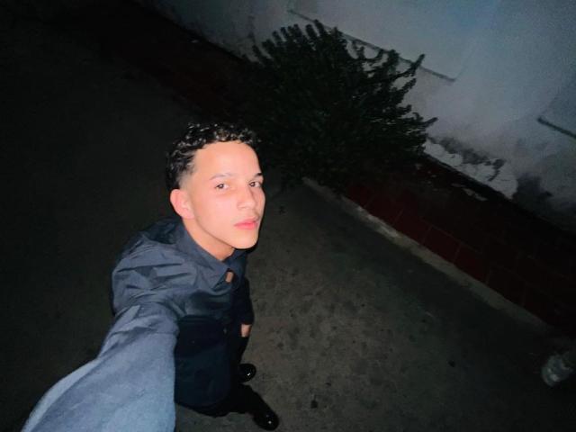 MidnightKing, a curly-haired dude with pale skin, takes a selfie from above in a dark, urban alley. He's wearing a gray shirt and black pants, looking down at the camera.